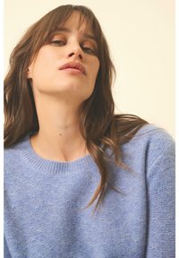 Hellblau gestrickter Pullover mit rundem Ausschnitt und strukturiertem Stoff. Das Modell hat welliges Haar, das teilweise die Schultern bedeckt.