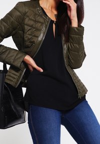 Olivenfarvet quiltet jakke med lynlås foran, ribbet krave og lange ærmer, parret med en sort top og blå jeans. Sort taske synlig.