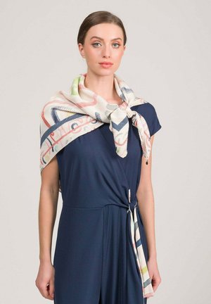 Donna che indossa un vestito blu navy con una sciarpa a motivo drappeggiata sulle spalle e annodata davanti, su uno sfondo semplice.