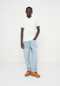 Λευκό πόλο πουκάμισο, ανοιχτό μπλε τζιν άνετης εφαρμογής και πορτοκαλί μπότες. Απλός σχεδιασμός με κοντά μανίκια, επίπεδη γιακά και ορατές λεπτομέρειες ραφής.