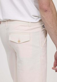 Pantalons en coton de couleur claire avec une poche arrière à fermeture à bouton. Le matériau a une texture lisse et un design ajusté.