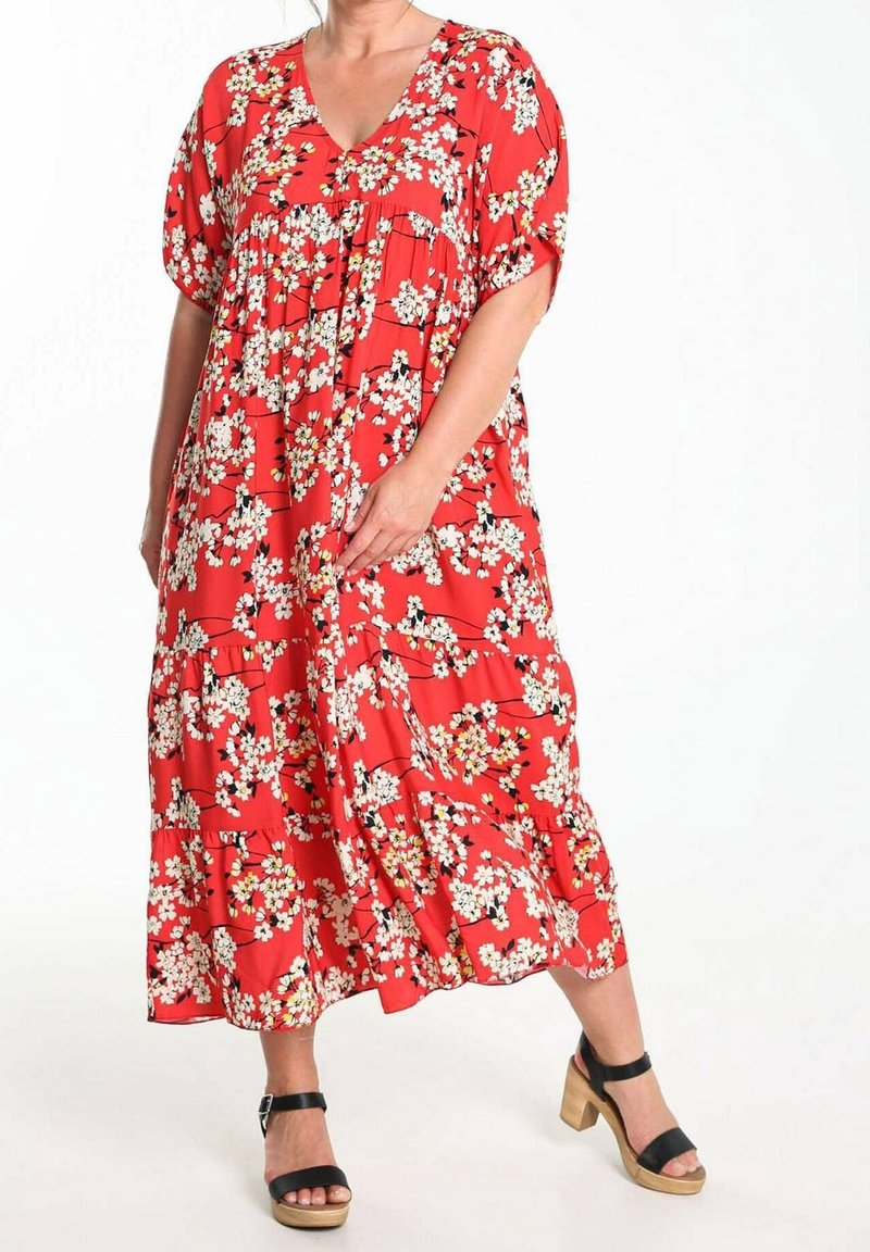 Robe maxi rouge fluide avec un décolleté en V, manches courtes, motif floral et ourlet à volants à étages. Portée avec des sandales noires à talons épais.