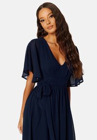 Námořnické modré šaty s průhlednými, lehce splývavými rukávy, výstřihem do V a páskem v pase. Materiál je hladký a splývavý, což podtrhuje siluetu.
