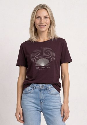 Uśmiechnięta blondynka, ubrana w ciemnofioletowy T-shirt z motywem muszli i napisem "Sun Sand Sea", w zestawieniu z jasnoniebieskimi dżinsami na jednolitym tle.
