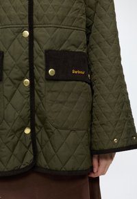 Veste matelassée Barbour vert olive avec surpiqûres losanges, boutons-pression dorés, bordure foncée et rabat de poche en velours côtelé avec broderie du logo jaune.