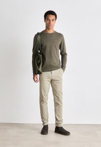 Maglione verde oliva con design a maglia testurizzata, pantaloni beige e stivali alla caviglia marroni scuri. La modella porta una borsa verde oliva.