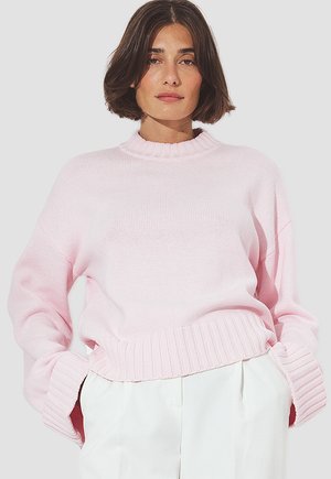 Woman with short brown hair wearing a light pink ribbed knit sweater and white high-waisted pants, posing with hands in pockets.
