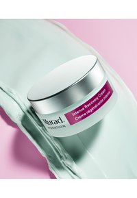 Intensiv återställningskräm i en klar burk med silverlock, med en lila etikett. Slät mynta-grön krämtextur på en rosa bakgrund.