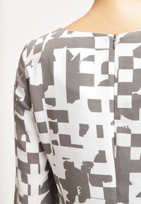 Pitkähihainen mekko, jossa on geometrinen harmaa ja valkoinen kuvio. Varustettu pyöreällä kaula-aukolla ja piilovetoketjulla takana. Sujuva kangasrakennett.