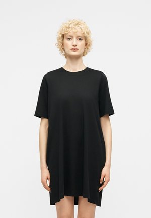 Fiatal személy rövid, göndör szőke hajjal, laza, fekete, rövid ujjú ruhát viselve, egyszínű, világos háttér előtt állva.