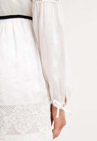 Vestido blanco con mangas largas y abullonadas, con intrincados detalles de encaje, una cintura texturizada y un cinturón negro como acento, con lazos en los puños.