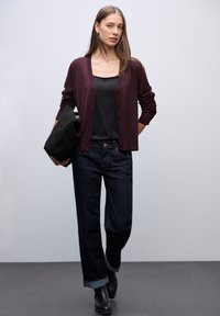 Cardigan bordeaux con bottoni, top nero, jeans in denim scuro con risvolti, e stivaletti neri alla caviglia, mentre tiene una borsa nera. Sfondo neutro.