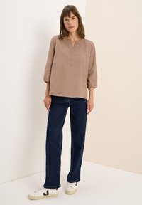 Femme debout portant un haut beige ample à manches trois-quarts, un jean bleu foncé et des baskets blanches avec un logo noir sur fond uni.