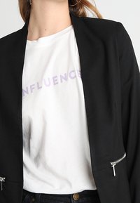 Musta räätälöity bleiseri avoimella etuosalla, jossa on hopeiset vetoketjutaskut, päällä valkoinen T-paita, jossa on violetilla painettu "INFLUENCE".