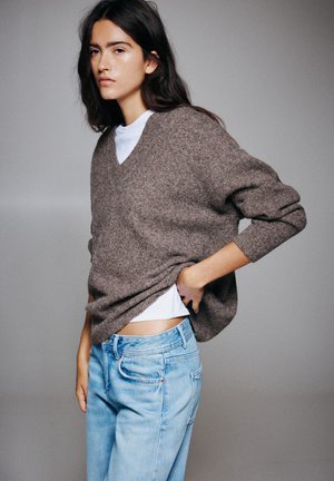 Un mannequin pose devant un fond de studio gris, vêtu d’un pull marron par-dessus une chemise blanche et d’un jean bleu clair.
