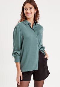 Chemise boutonnée vert clair avec col, présentant des manches longues et une coupe décontractée, associée à un short noir. Texture lisse.