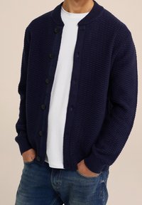 Cardigan texturé bleu marine avec col rond, fermeture boutonnée devant, poignets côtelés, et un t-shirt blanc lisse en dessous, associé à un jean bleu.