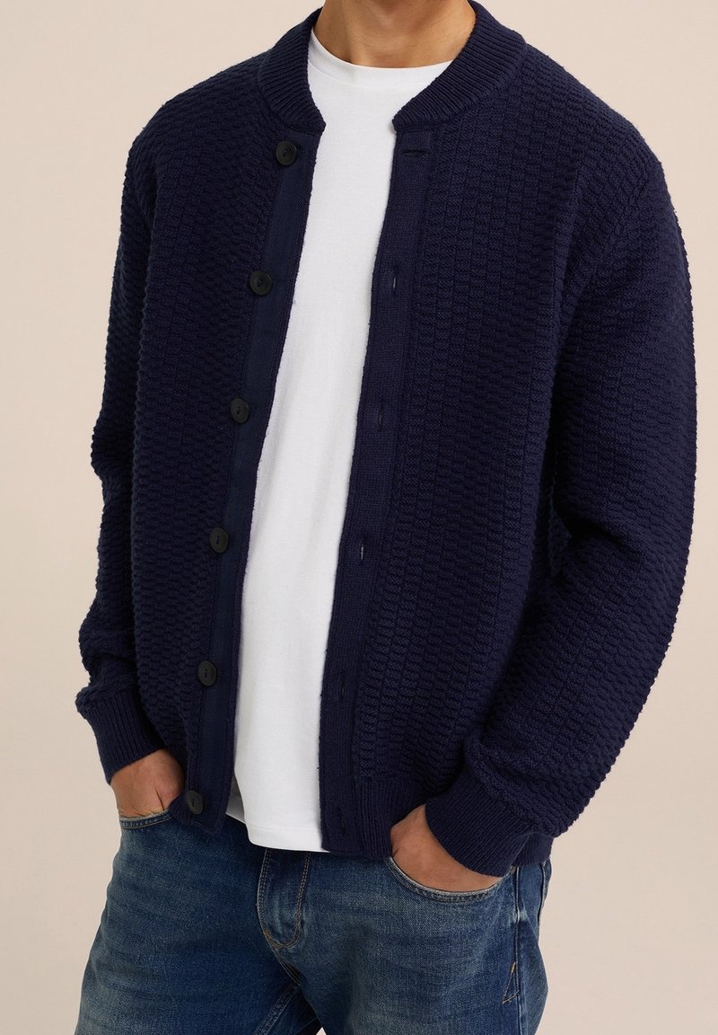 Cardigan texturé bleu marine avec col rond, fermeture boutonnée devant, poignets côtelés, et un t-shirt blanc lisse en dessous, associé à un jean bleu.