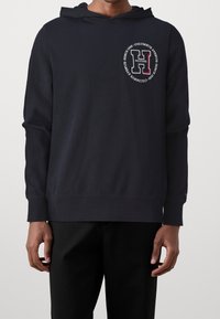 Námořnická mikina s kapucí, s kulatým grafickým logem v červené a bílé barvě na prsou. Dlouhé rukávy a pružné manžety, jemný materiál.