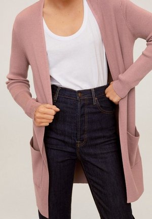 Vrouw draagt een stoffig roze lange cardigan over een wit t-shirt met ronde hals en donkerblauwe hoog getailleerde jeans, handen houden de randen van de cardigan vast.