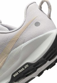 Lys grå atletiske sko med textured mesh øverdel. Har en beige swoosh, lille grå prik, polstret krave og en sort skridsikker sål.