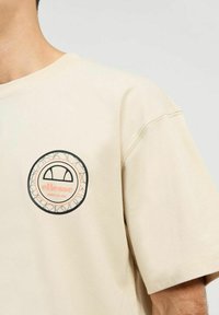 Camiseta de algodón beige con un logo gráfico circular en negro y rosa en el pecho, de corte relajado y mangas cortas.