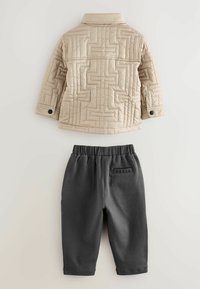 Beige quiltet jakke med geometrisk syning og sorte knapper, kombineret med mørkegrå bukser med elastisk talje, mærket "BAKER" på ryggen.