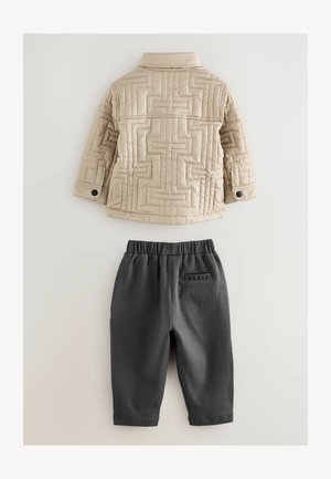 Beige gewatteerde jas met geometrische stiksels en zwarte drukknoopjes, gecombineerd met donkergrijze broek met elastische tailleband, gemarkeerd met "BAKER" op de achterkant.