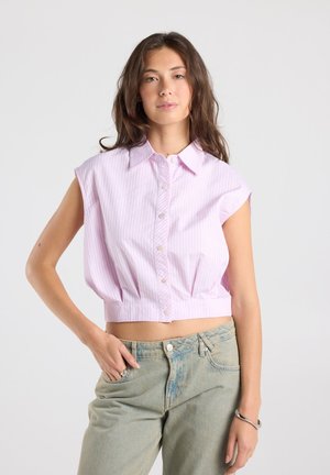 Jeune femme aux longs cheveux portant un haut court sans manches rose rayé et un jean bleu clair, posant avec une main dans la poche.