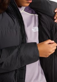 Osoba ubrana w fioletową bluzę The North Face, otwierająca czarną pikowaną kurtkę, skupiając się na wewnętrznej kieszeni kurtki zapinanej na zamek.