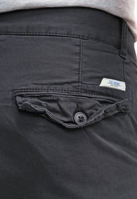 Pantalones negros con un bolsillo con tapa abotonada, hechos de una tela resistente. Se puede ver una pequeña etiqueta con "EDC BRAND" cerca de la cinturilla.