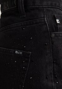 Mustad denim teksad, millel on tekstureeritud pind ja laiali jaotatud mustade helmeste kaunistused ning brändi silt tagakotil.