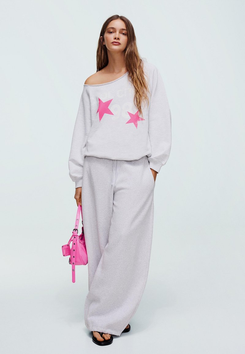 Vrouw draagt een witte off-shoulder sweatshirt met roze sterren, brede witte broek, zwarte sandalen, en houdt een kleine roze handtas vast.