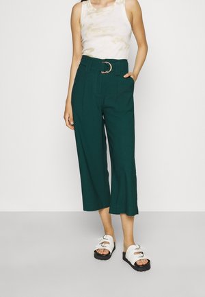 Grüne Culottes mit hoher Taille und weitem Bein, ausgestattet mit einem Gürtel mit einer silbernen runden Schnalle. Kombiniert mit einem beigen Batik-Tanktop und weißen Sandalen.