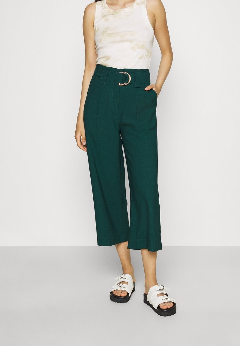 Gröna culotter med hög midja och vida ben, featuring ett bälte med ett silverfärgat runt spänne. I kombination med en beige tie-dye linne och vita sandaler.