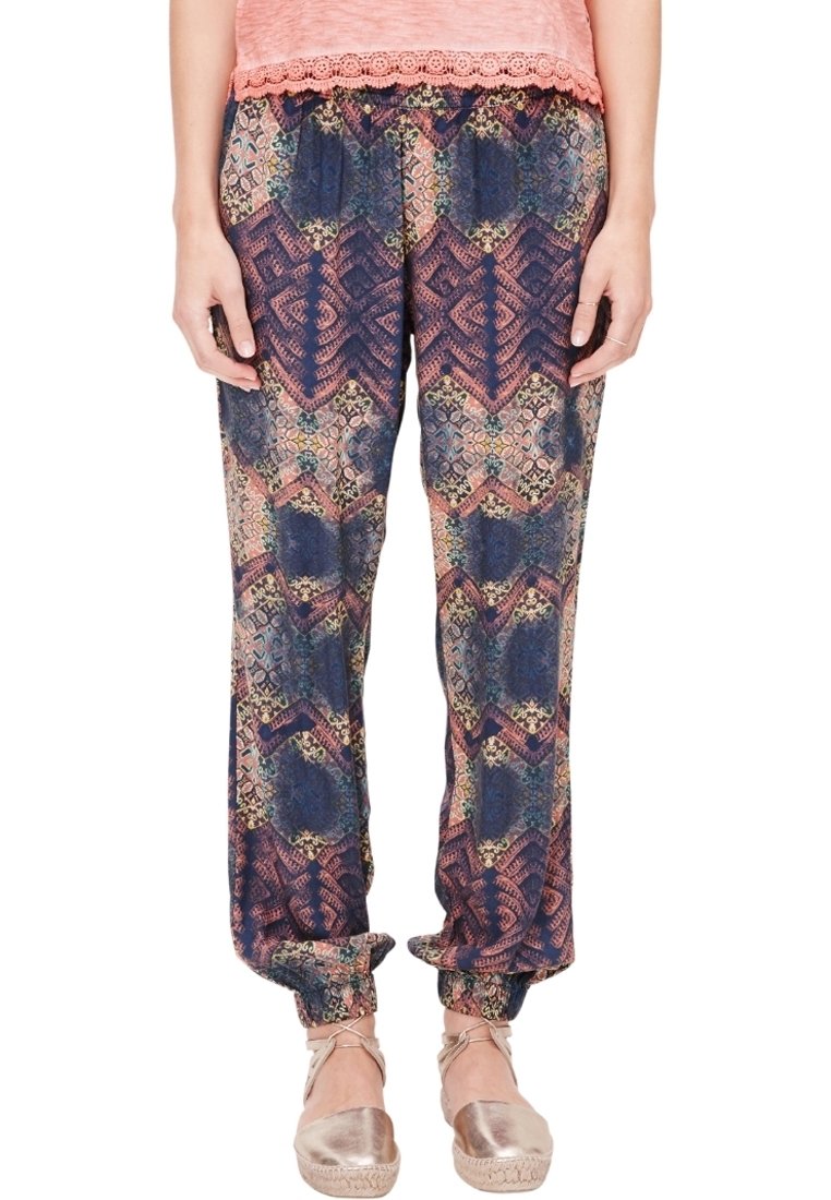 Femme portant un pantalon ample à motif géométrique bleu marine et rose, un haut pêche orné de dentelle et des espadrilles beiges, debout, les mains le long du corps.
