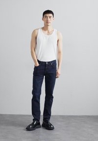 Top blanco de tirantes, jeans oscuros y zapatos negros. El modelo se encuentra de pie contra un fondo neutro, enfatizando el ajuste y el color de la ropa.