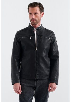 Mand iført sort læderjakke med lynlås og to brystlommer med lynlås og mørke jeans, stående mod en ensfarvet lys baggrund.