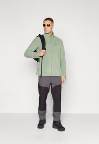 Lichtgroene fleecejack met rits, donkergrijze en zwarte broek, zwarte rugzak en zonnebril. Gestructureerd materiaal met een relaxte pasvorm.