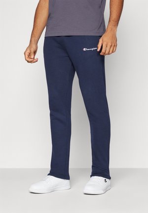 Man draagt marineblauwe Champion joggingbroek en witte sneakers, staand tegen een effen lichte achtergrond.