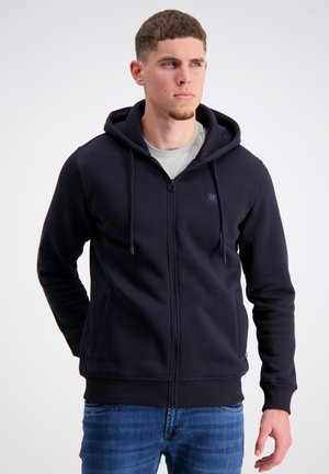 Mørkeblå zip-up hoodie med snørehætte, frontlommer, ribbede manchetter og kant. Glat tekstur, båret over en lys grå skjorte.