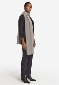 Lys grå tørklæde lavet af blødt materiale, draperet over en sort top. Modellen har sorte tætsiddende bukser og spidse sorte ankelstøvler på.