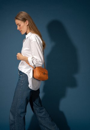 Donna con capelli lunghi che indossa una camicia bianca e jeans blu, che cammina con una piccola borsa a tracolla in pelle marrone su sfondo blu scuro.