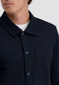 Marineblaues Hemdjackett mit knöpfbarem Frontverschluss, ausgestattet mit einem Kentkragen und einer Brusttasche. Der Stoff hat ein dezentes Rippmuster.