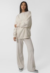 Kreem värvi üleliigne kapuutsiga särk ribitud varrukatega, koos heledate beežide laia jala pükstega. Mõlemad on valmistatud pehmetest materjalidest, millel on peened õmbluse detailid.