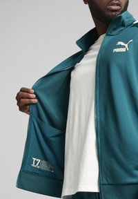 Teal zip-up jacka i mjukt material med Puma-logotypen på bröstet och en etikett inuti. Bärs över en vit T-shirt.