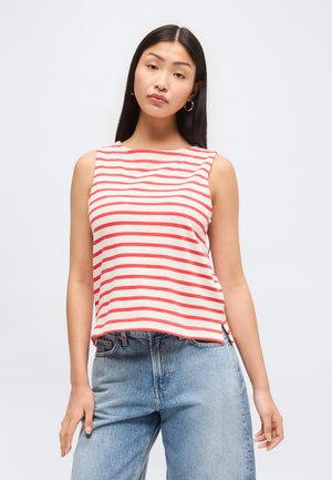 Kobieta z długimi ciemnymi włosami nosi złote kolczyki koła, bezrękawnik w czerwono-białe paski i jasnoniebieskie dżinsy, stojąc na jednolitym tle.