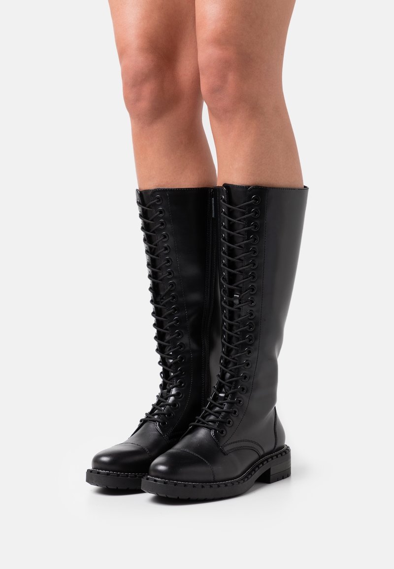 Bottes noires montantes à lacets avec fermeture éclair latérale, portées sur des jambes nues, debout sur une surface blanche.