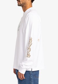 Witte lange mouw shirt met zijsplitten, met een groene graphic op de voorkant en bruine "RVCA" tekst langs de linker mouw.