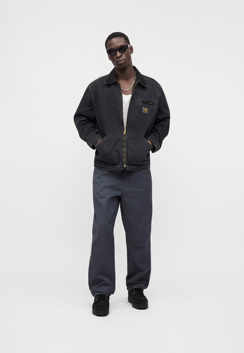 Mann mit schwarzen Sonnenbrillen, schwarzer Zip-up-Jacke, weißem Hemd, grauer Hose und schwarzen Schuhen steht mit den Händen in den Taschen vor schlichtem Hintergrund.